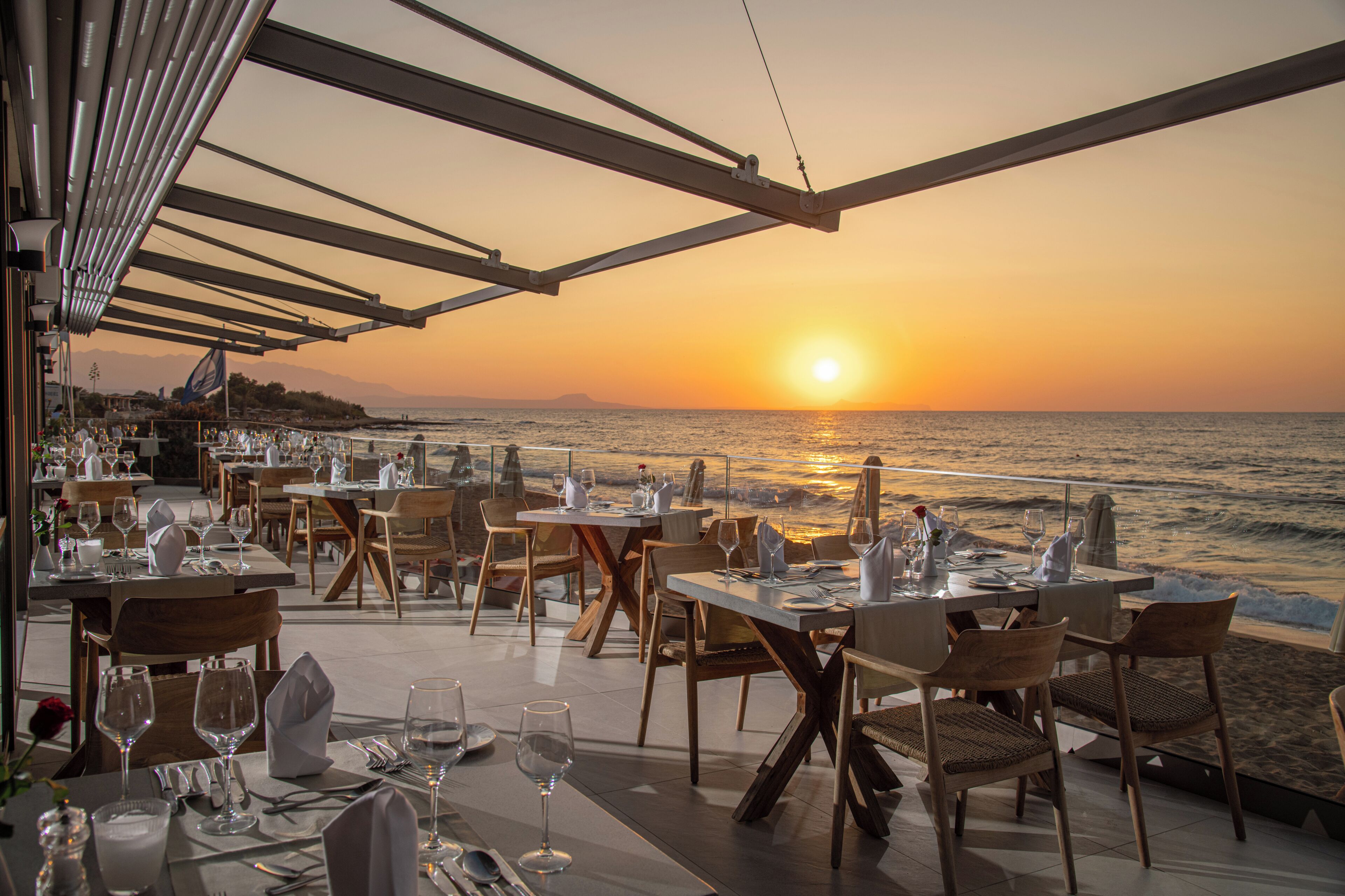 An outdoor dining area with tables and chairs facing the ocean at sunset.