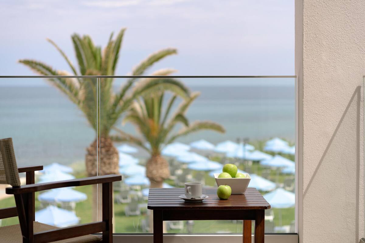 Uma varanda com vista para o oceano, uma mesa com uma tigela de maçãs verdes, uma xícara e uma cadeira.