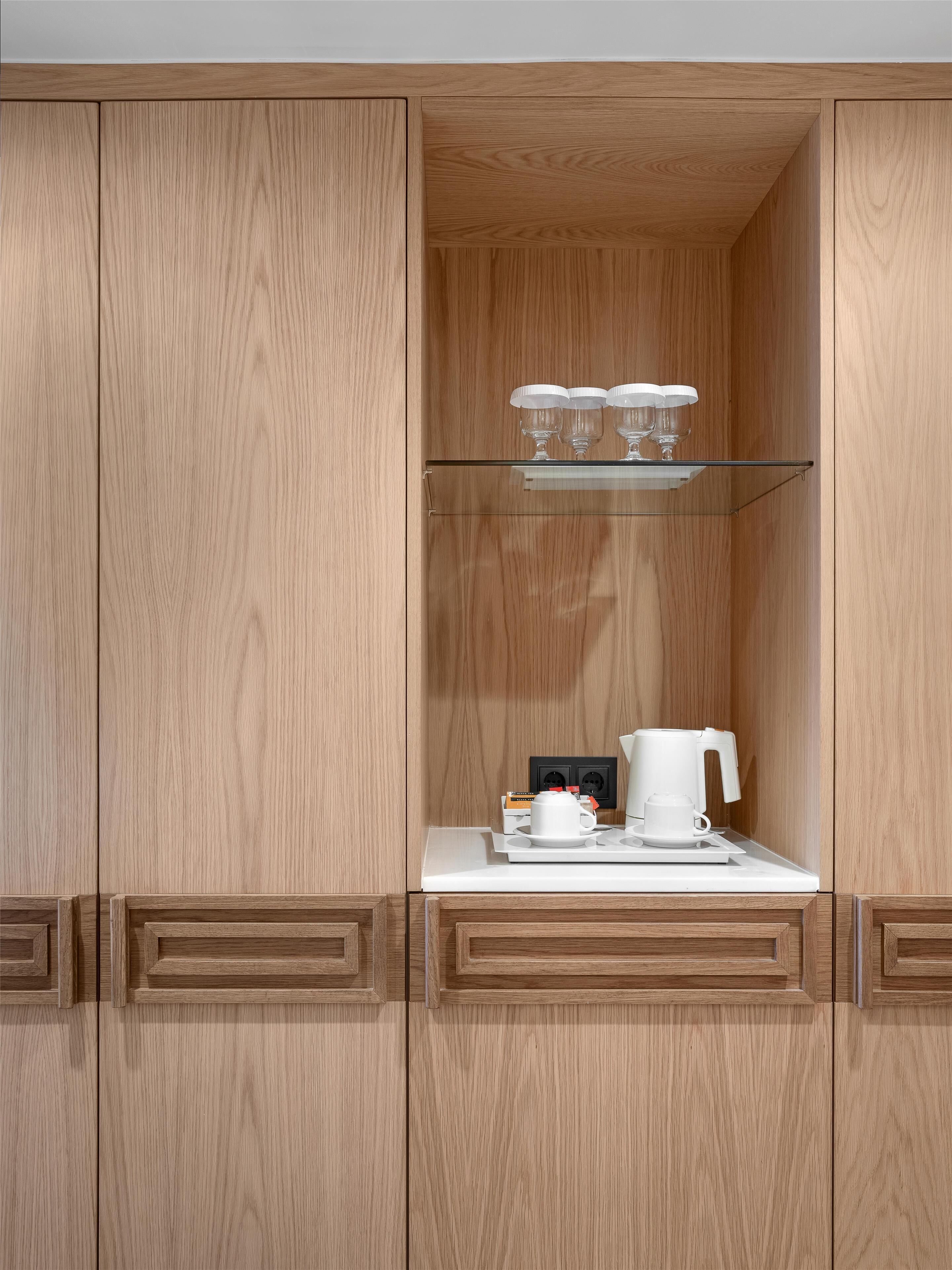 A wooden cabinet with a glass shelf holding three glasses and a white coffee maker on a white countertop.