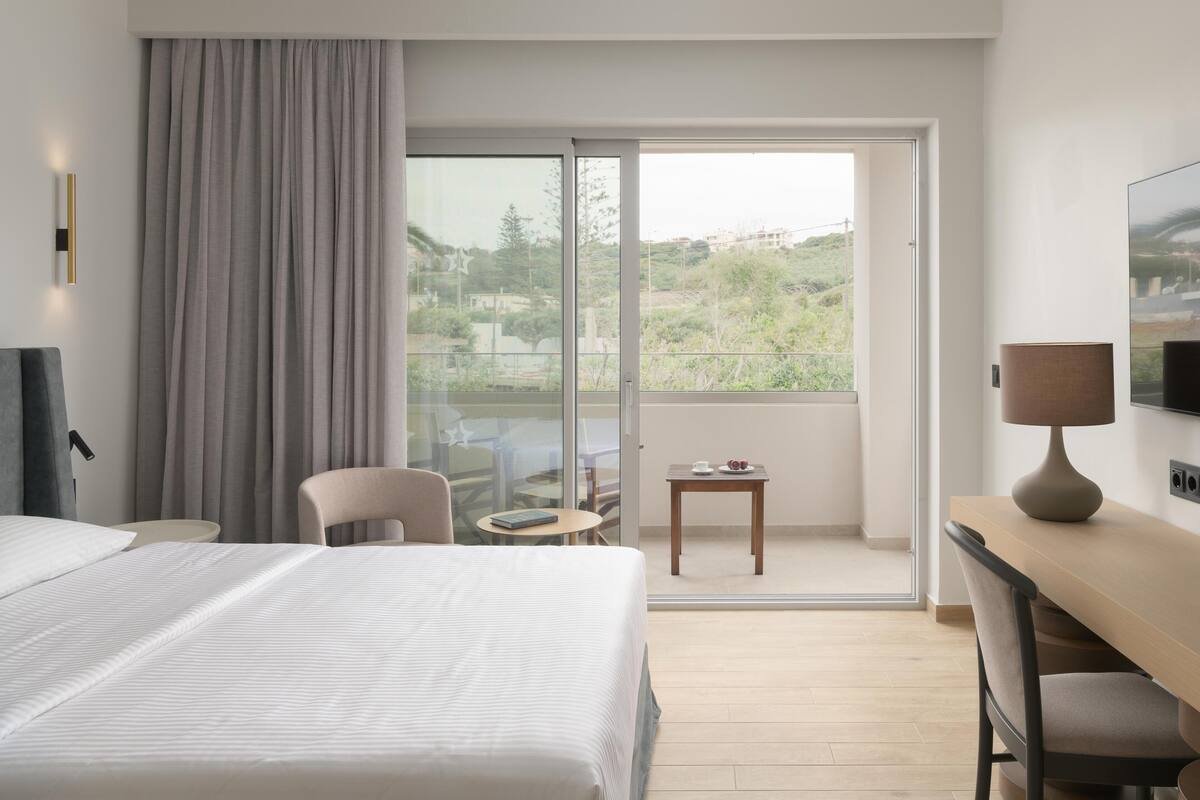 Quarto de hotel com uma cama grande, uma escrivaninha, uma cadeira, uma pequena mesa com uma xícara e um livro, e uma varanda com vista para áreas verdes e edifícios.