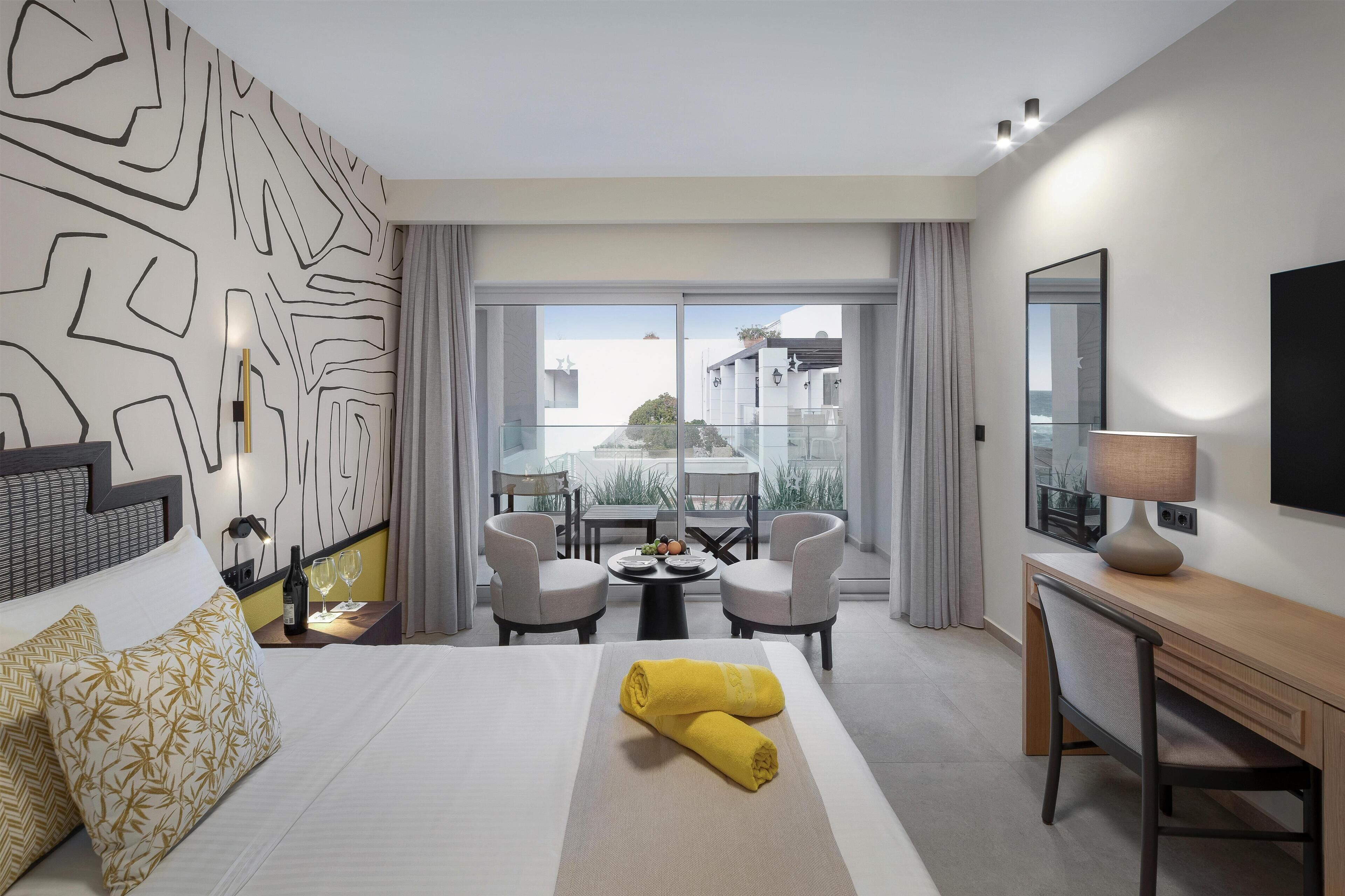 A hotel room with a bed, a desk, a chair, a table with a bottle and glasses, and a view of the outside.