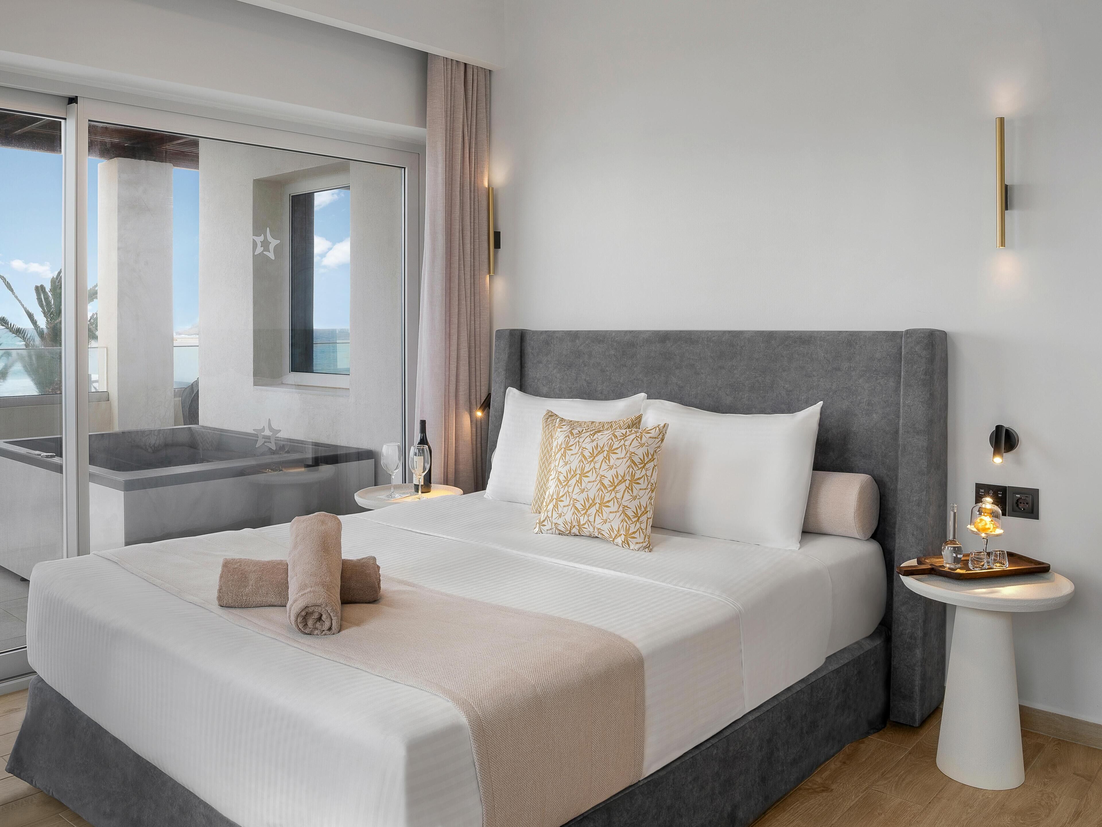A modern hotel room with a large bed, a bedside table with a lamp, a bathroom with a glass shower, and a view of the ocean.