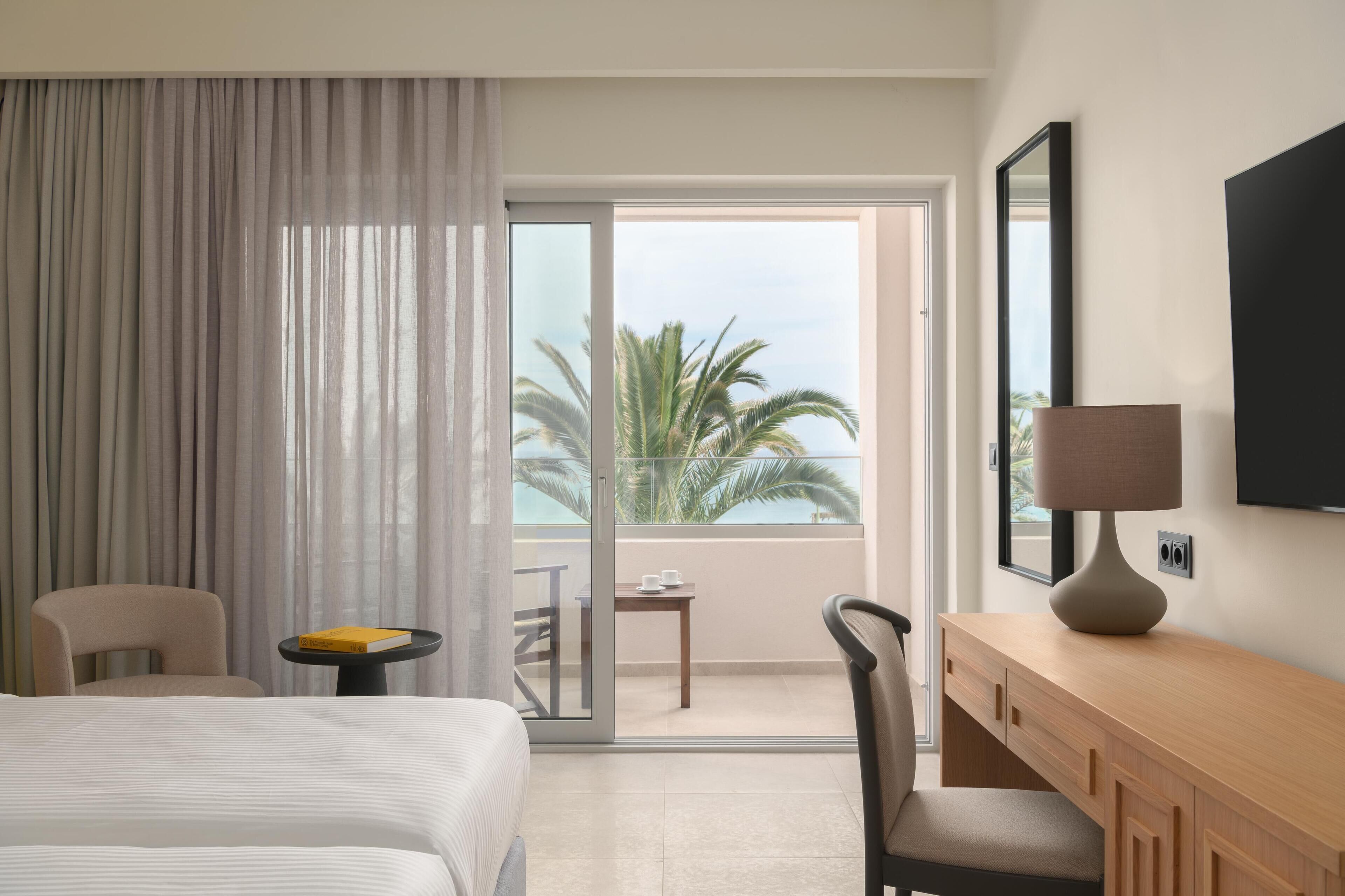 Habitación de hotel con cama, escritorio, silla, una mesa con un libro y vista a palmeras a través de una puerta corrediza de vidrio.