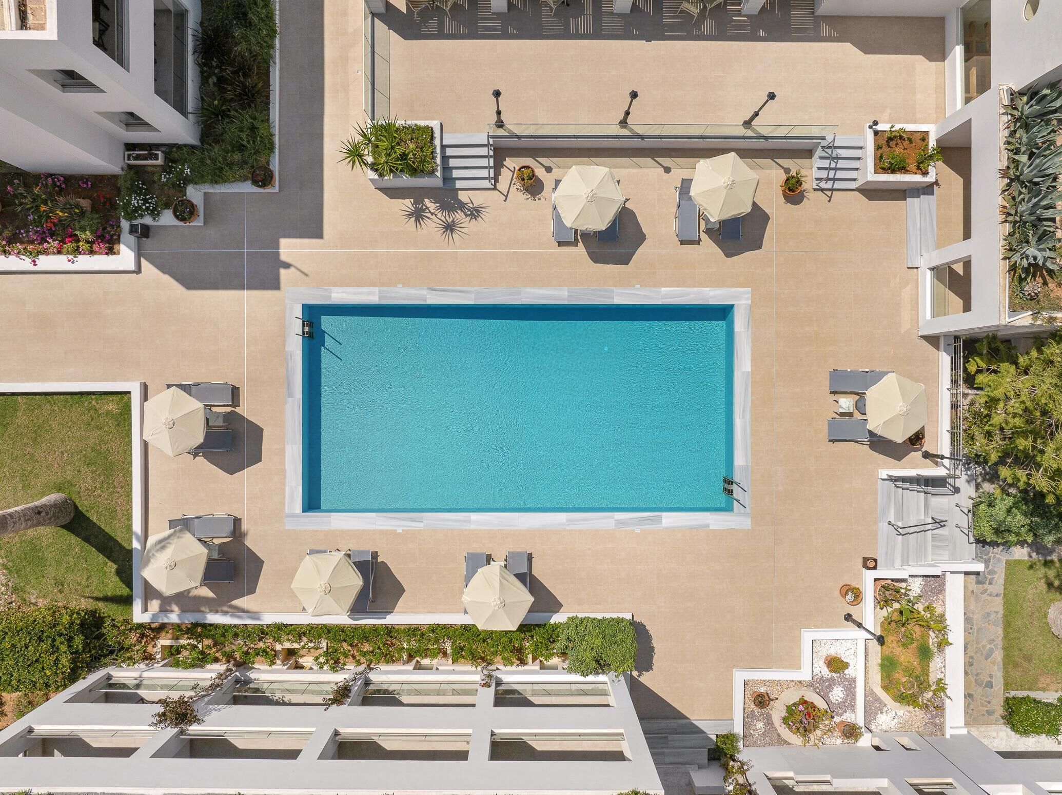 An aerial view of a rooftop pool area with a rectangular pool, surrounded by lounge chairs and umbrellas, and bordered by potted plants and greenery.