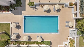 An aerial view of a rooftop pool area with a rectangular pool, surrounded by lounge chairs and umbrellas, and bordered by potted plants and greenery.