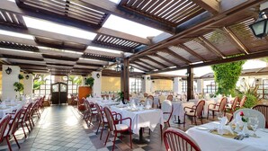 Amplia zona de comedor al aire libre con varias mesas dispuestas para comer, rodeada de sillas rojas, bajo una pérgola de madera con luz natural.