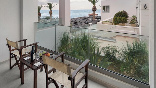 A balcony with a table and chairs, overlooking a coastal area with palm trees and a view of the sea. - Creta Royal - Adults only (Rethymno)