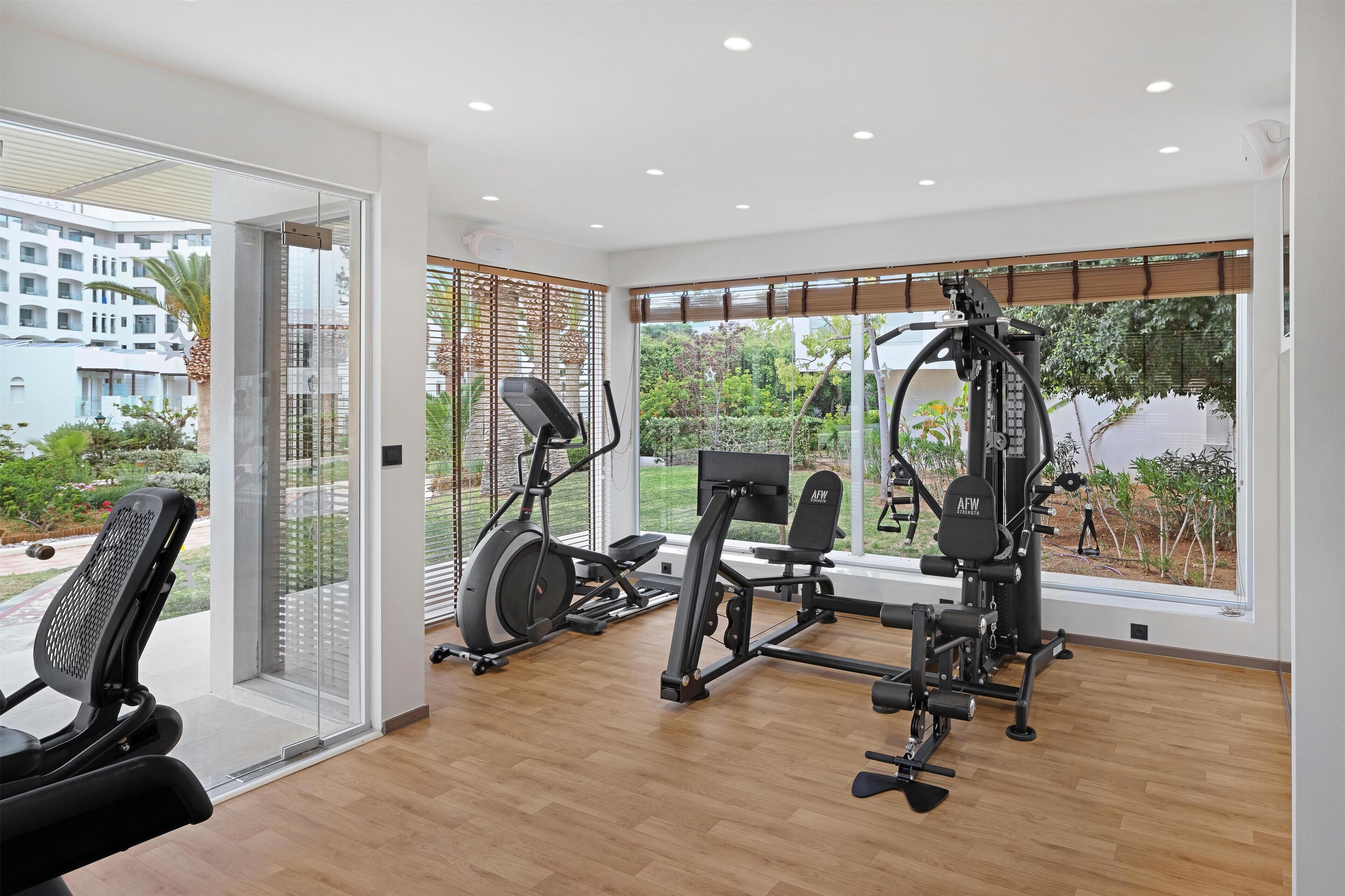 A modern gym with cardio machines and free weights, large windows with a view of greenery and buildings.