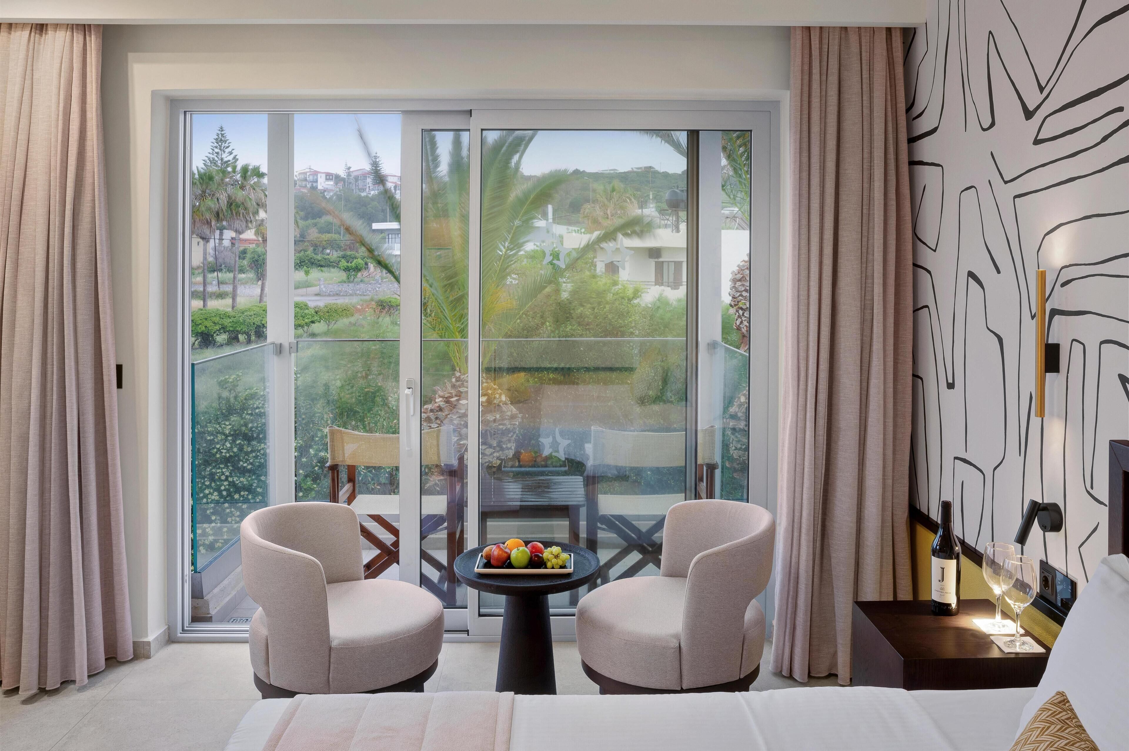 A hotel room with a balcony, two armchairs, a small table with fruit, a bottle of wine, and a bed.