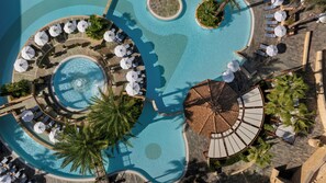 A large, circular swimming pool with multiple sun loungers and umbrellas, surrounded by palm trees and a stone deck.