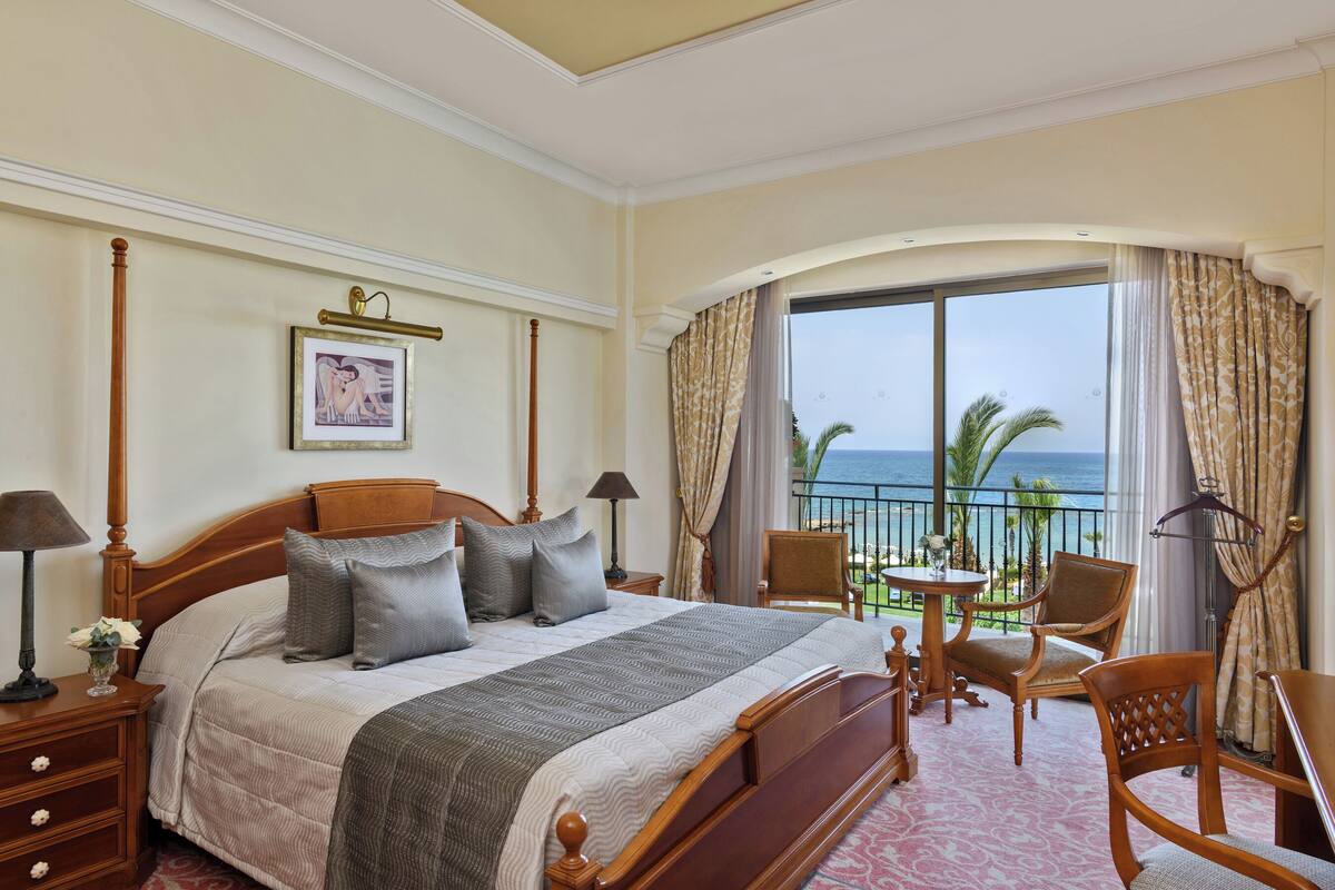 A hotel room with a large bed, a balcony with a view of the ocean, a small table with chairs, and a framed picture on the wall.