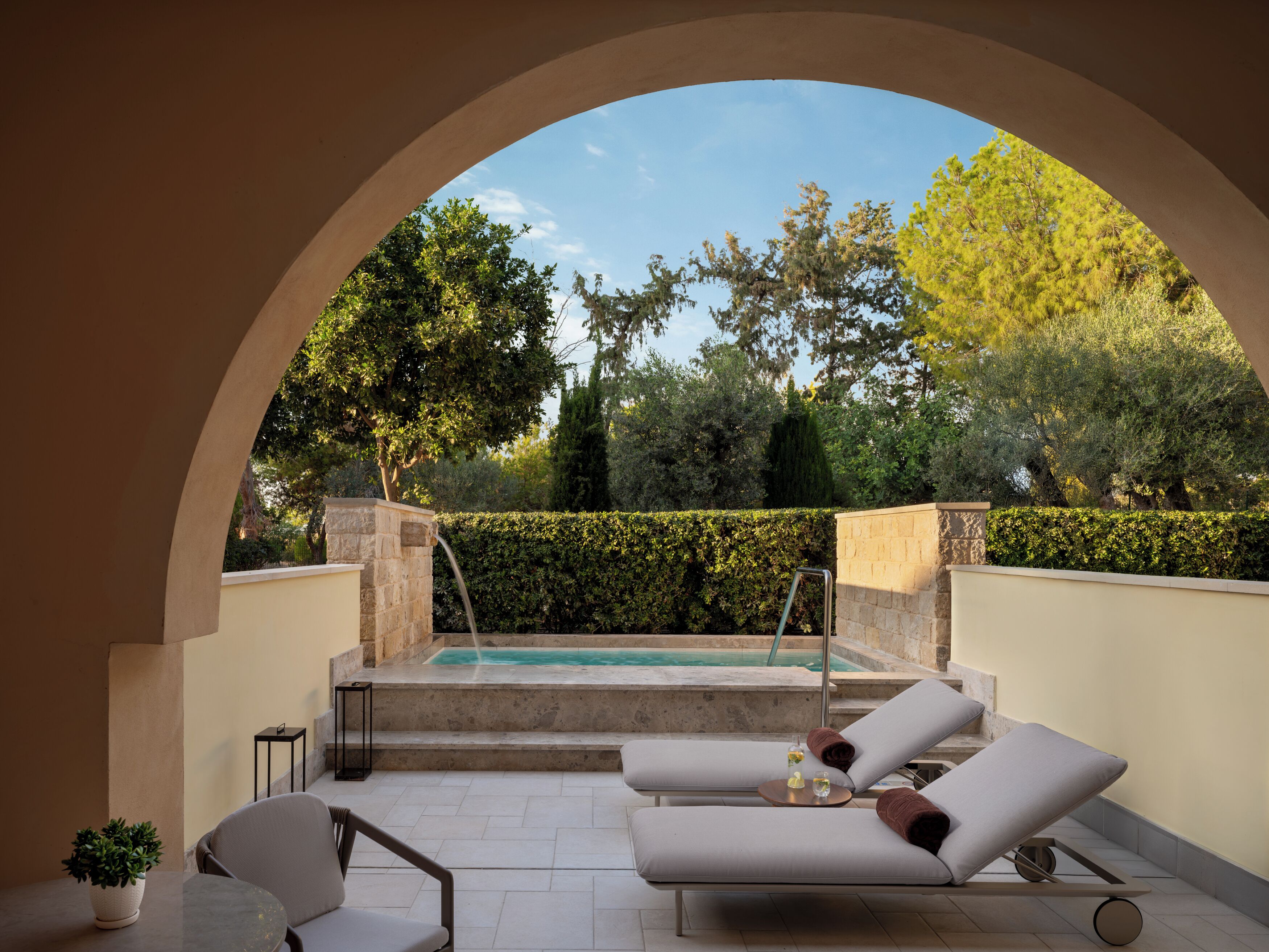 A sunlit outdoor lounge area with a pool, two lounge chairs, a small table, and a potted plant.