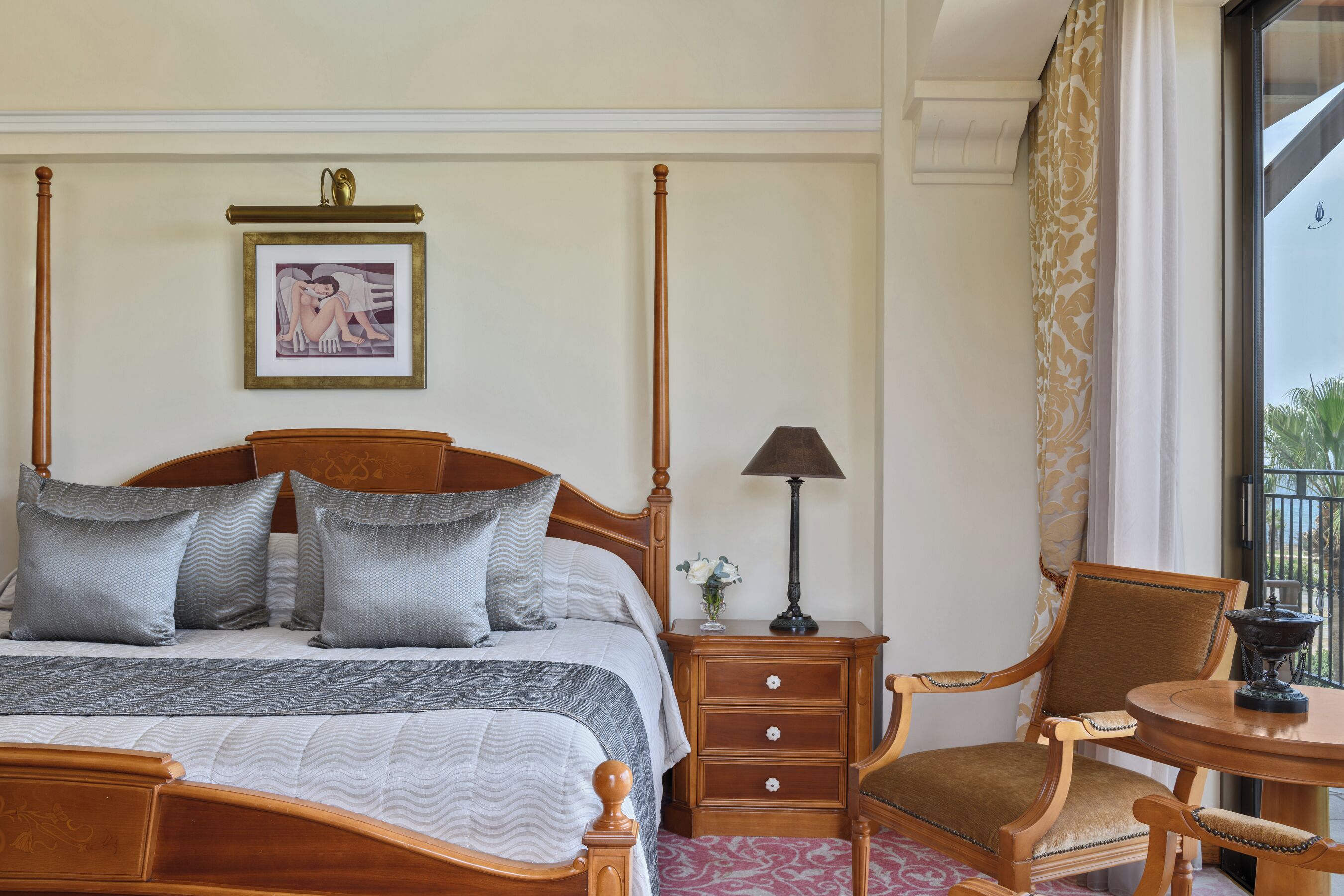 Una habitación de hotel con una cama con dosel, una mesita de noche con lámpara, una silla, una pequeña mesa y un cuadro en la pared.