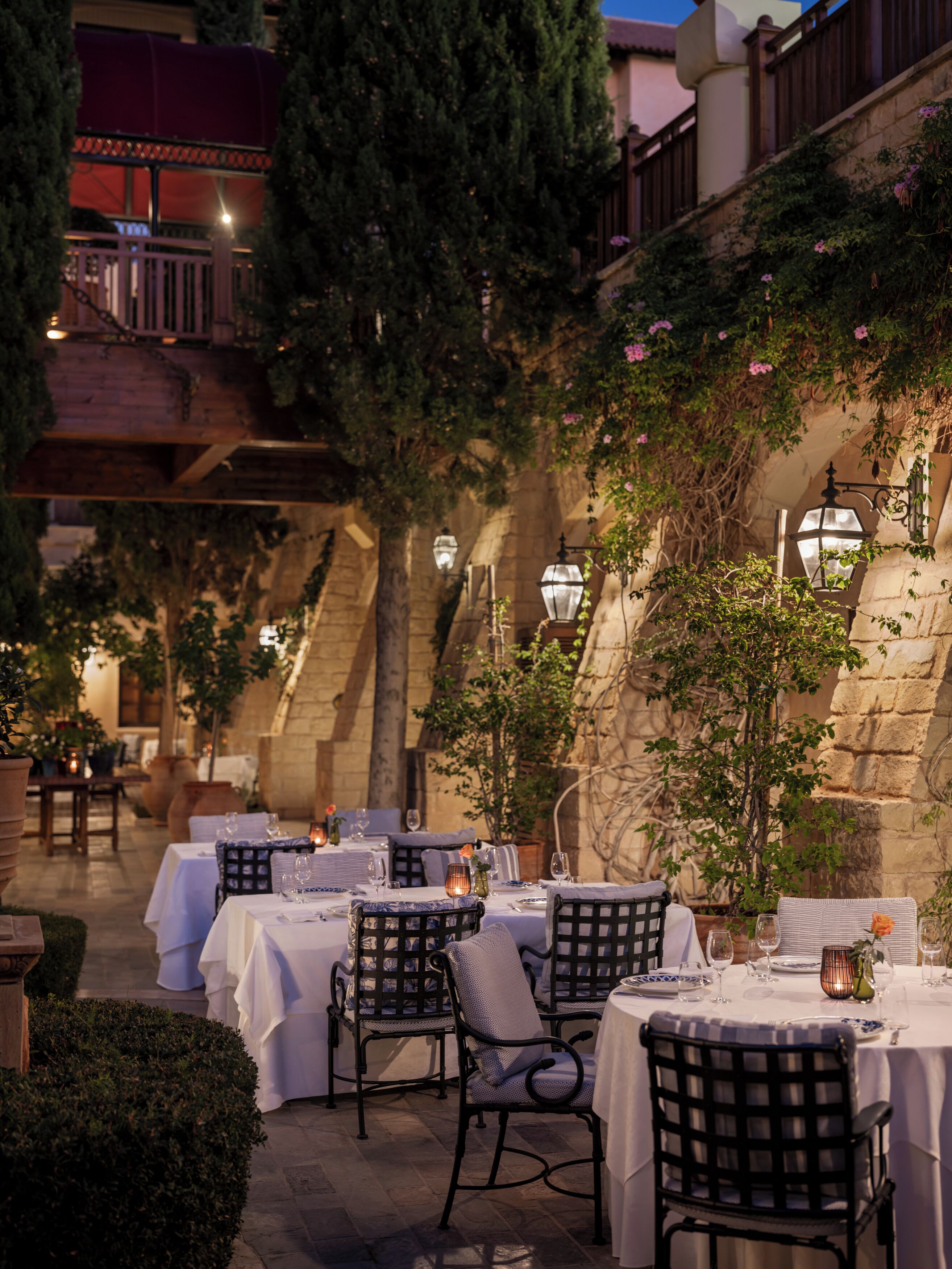 An outdoor dining area with tables set for two, surrounded by stone walls and greenery.