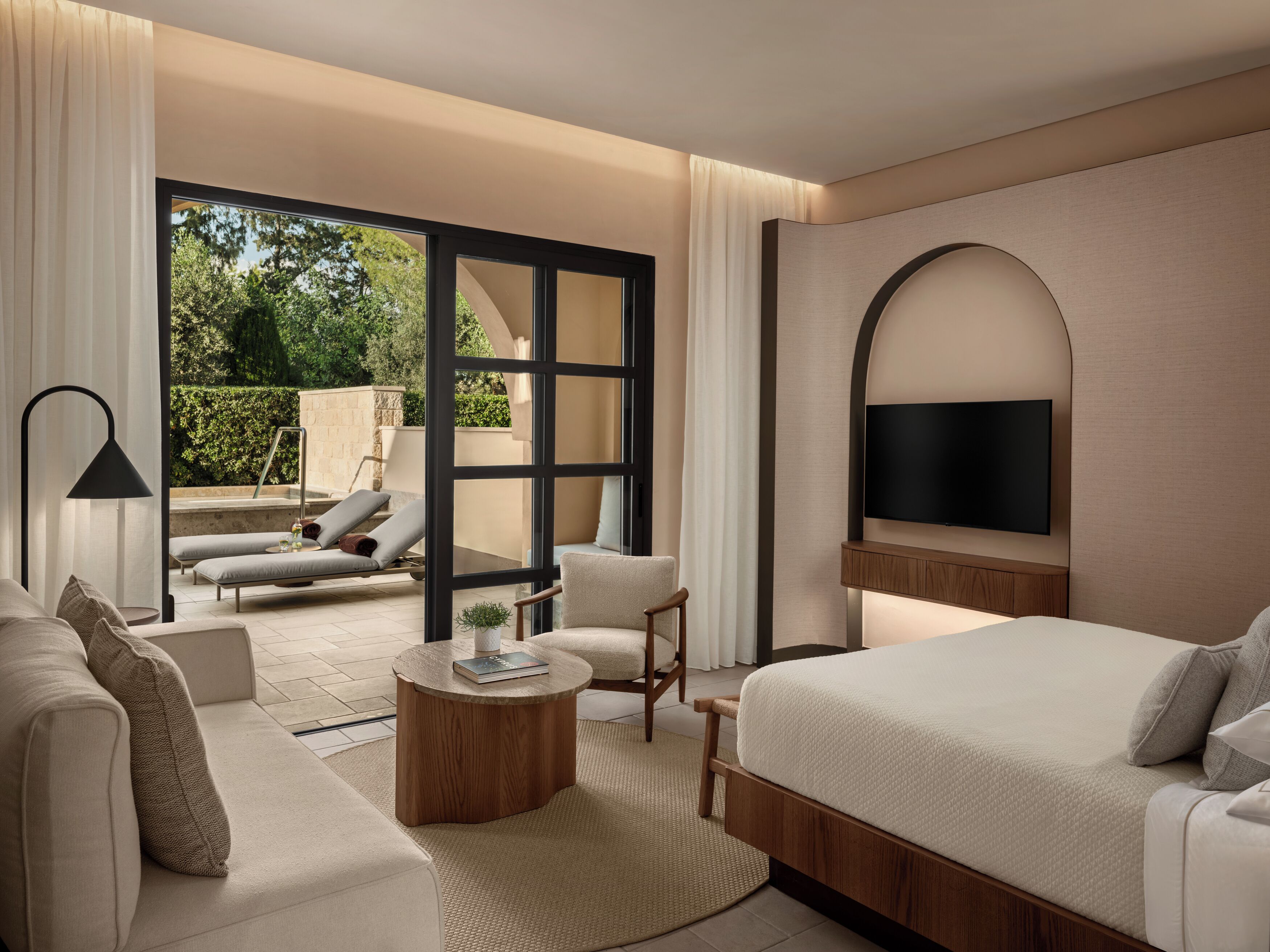 A modern hotel room with a bed, sofa, TV, and a view of the outdoors.