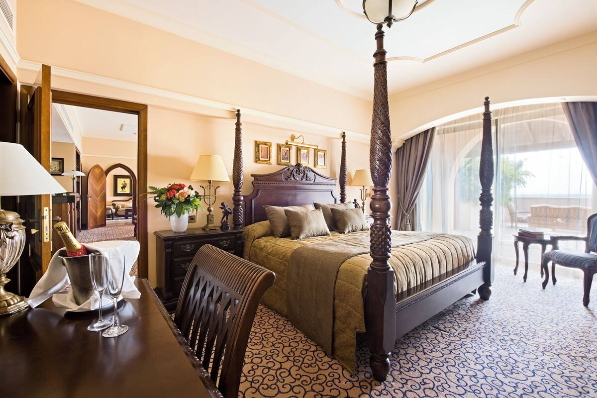 A four-poster bed with a canopy, a chandelier, a dining table with a vase and champagne glasses, and a view of the outdoors through large windows.
