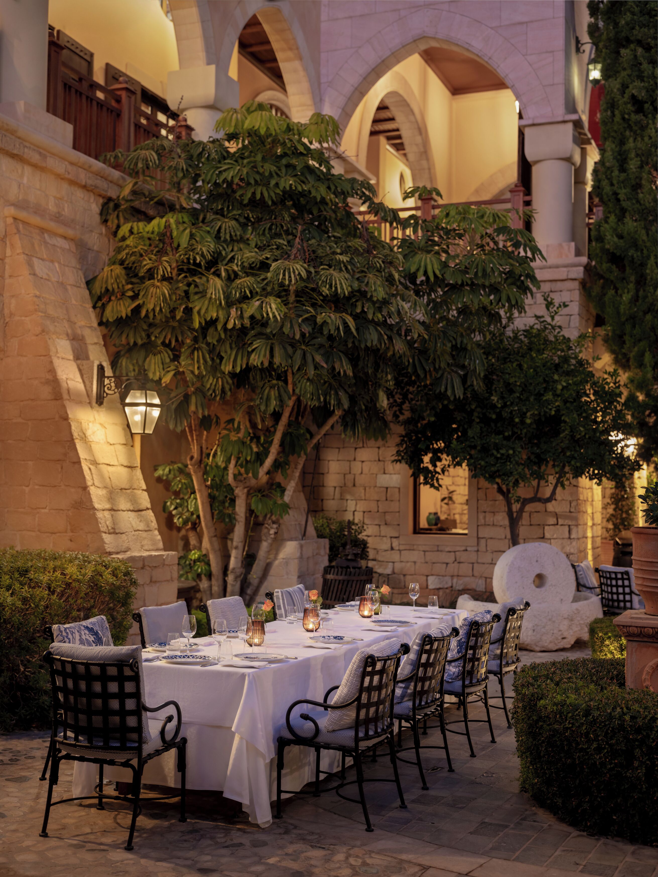 An outdoor dining area with tables set for two, surrounded by stone walls and greenery.