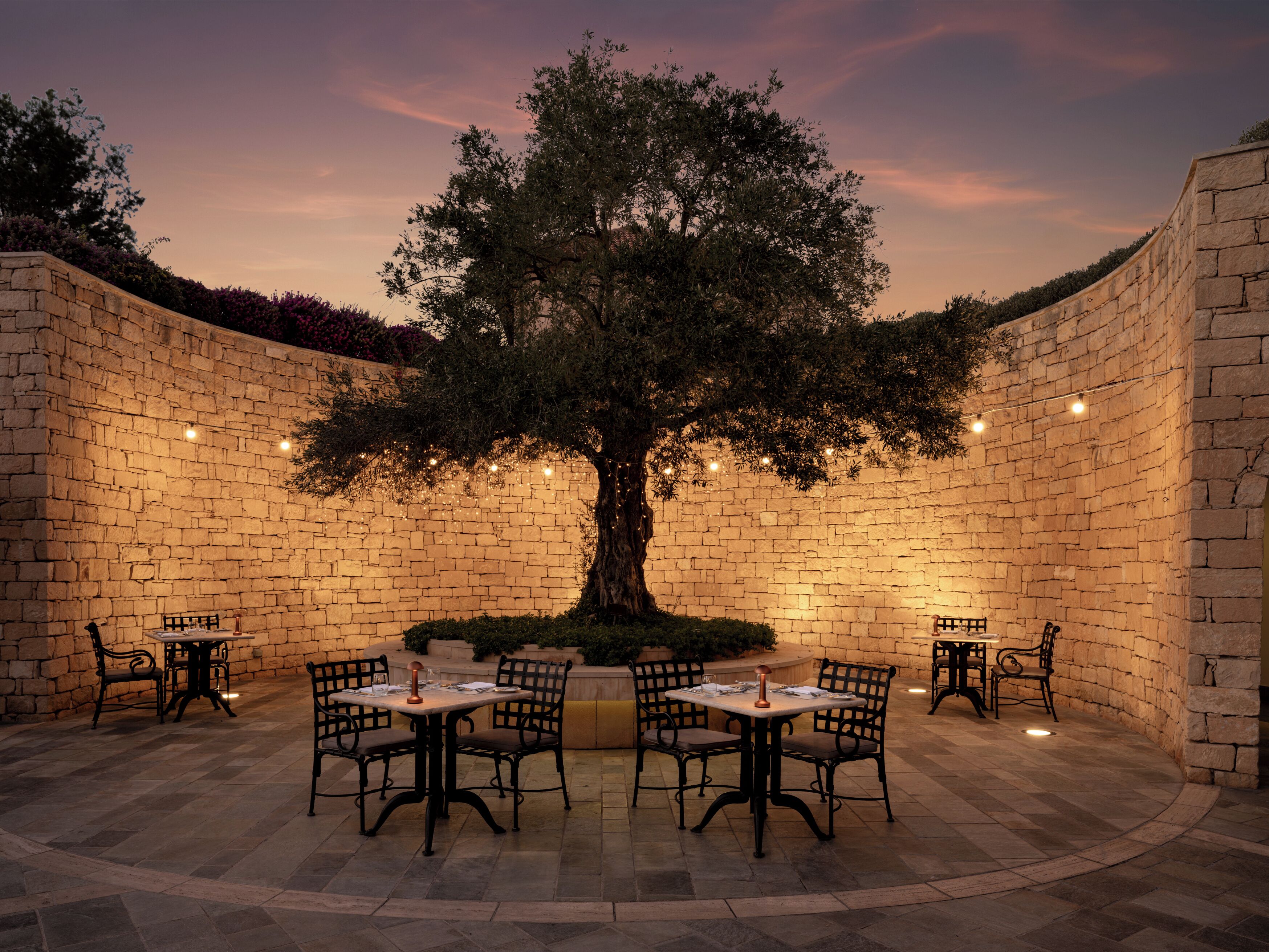 Un espace extérieur aménagé avec des murs en pierre, comprenant un arbre central, des tables et des chaises.
