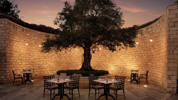 A stone-walled outdoor seating area with a central tree, tables, and chairs.