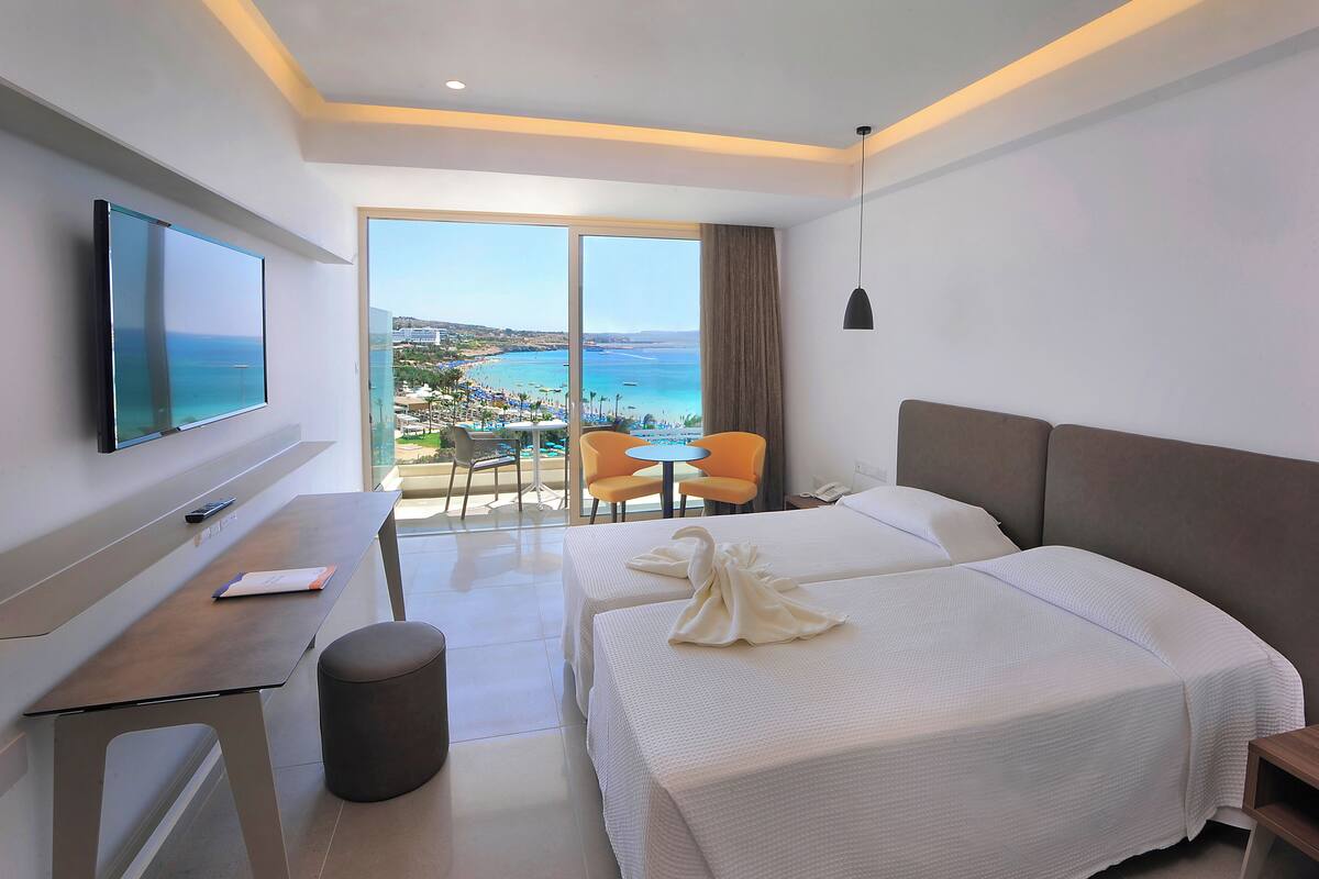 Une chambre d’hôtel avec deux lits, un bureau, une télévision et un balcon offrant une vue sur la mer et la plage.