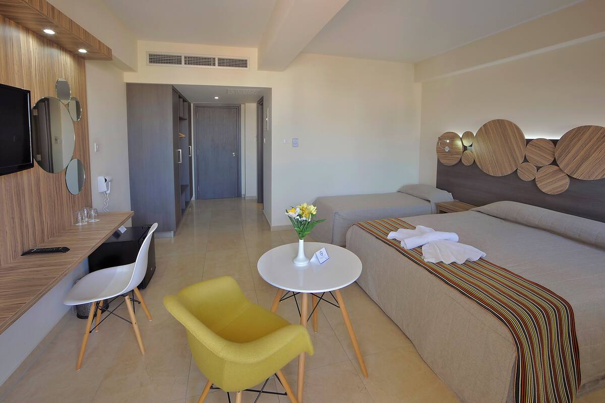 Une chambre d’hôtel moderne avec un canapé, un fauteuil jaune, une petite table avec un vase et un téléviseur fixé au mur.