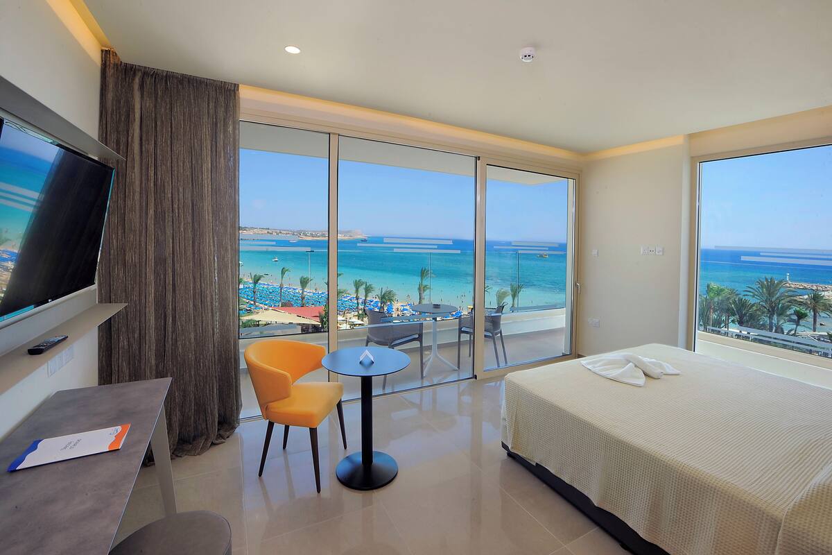 Une chambre d’hôtel comprenant un lit, un bureau, une chaise, une petite table et un téléviseur. La chambre dispose de grandes fenêtres offrant une vue sur la mer et la plage.