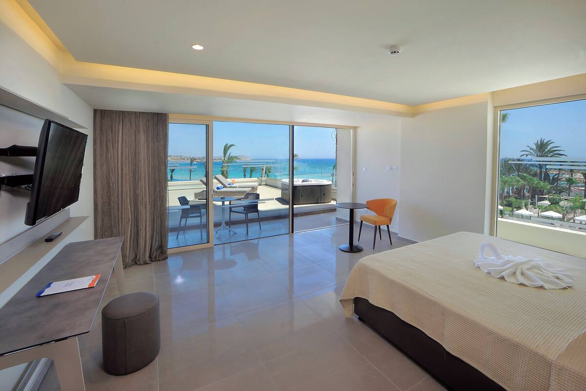 Une chambre d’hôtel avec un lit, un bureau, une télévision, un balcon donnant sur une piscine et agrémenté de chaises, et une vue sur l’océan.