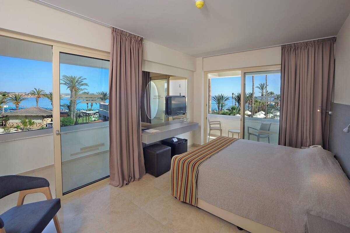 Une chambre d’hôtel avec un lit, un bureau avec une télévision, une chaise et un balcon offrant une vue sur la plage et les palmiers.