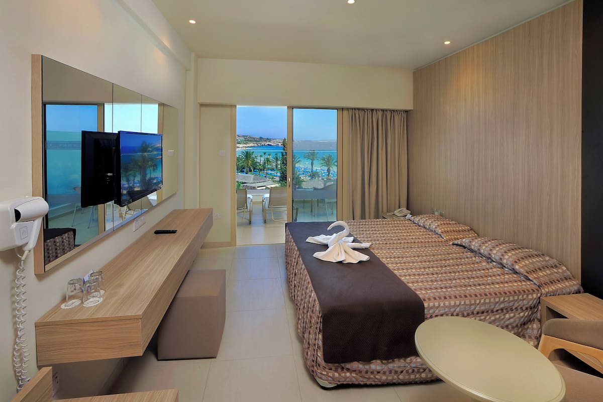 Une chambre d’hôtel dotée d’un grand lit, d’un bureau avec un téléviseur à écran plat, d’un sèche-cheveux et offrant une vue sur la plage et les palmiers.