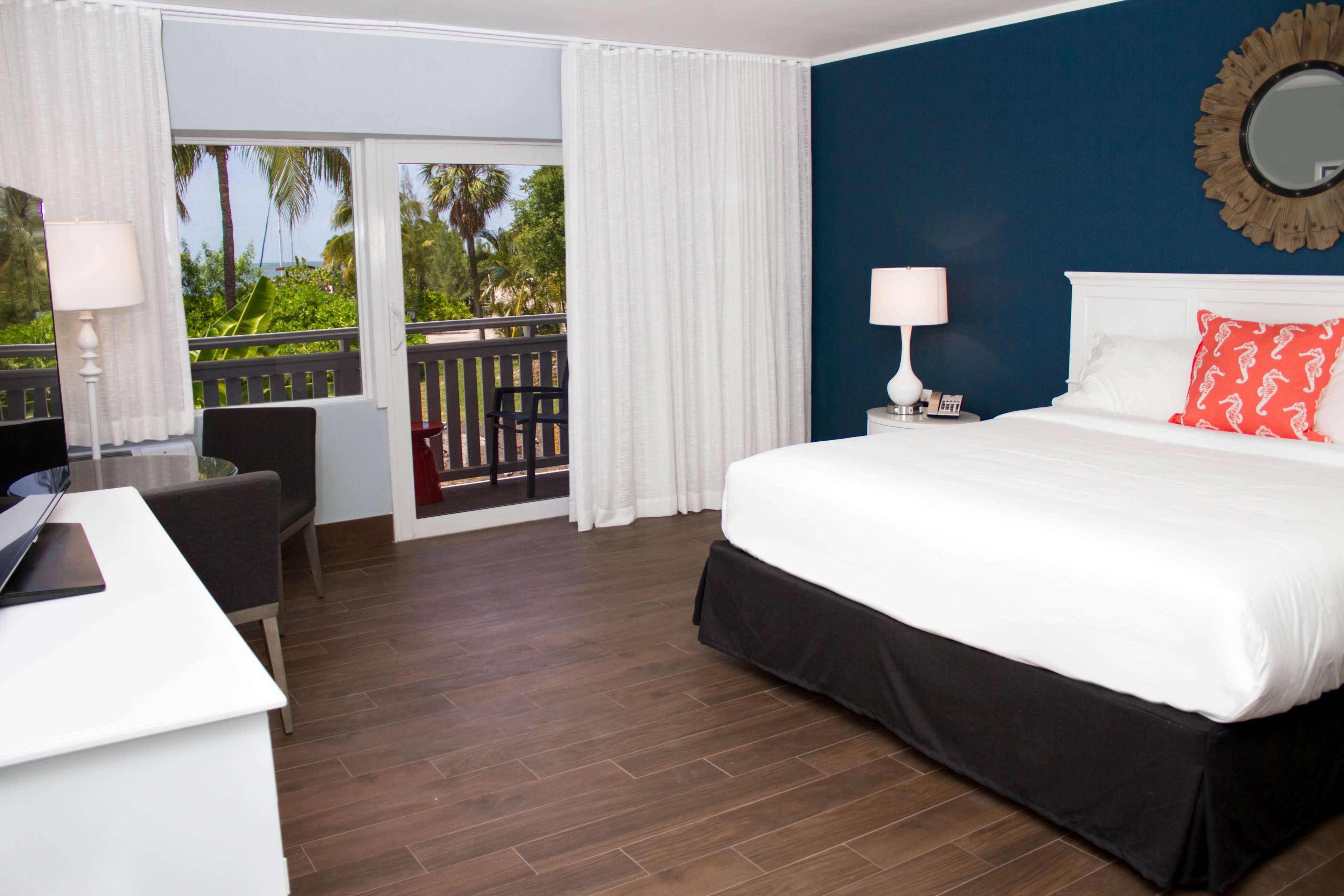 A hotel room with a bed, a desk, a chair, a balcony with curtains, and a view of palm trees.