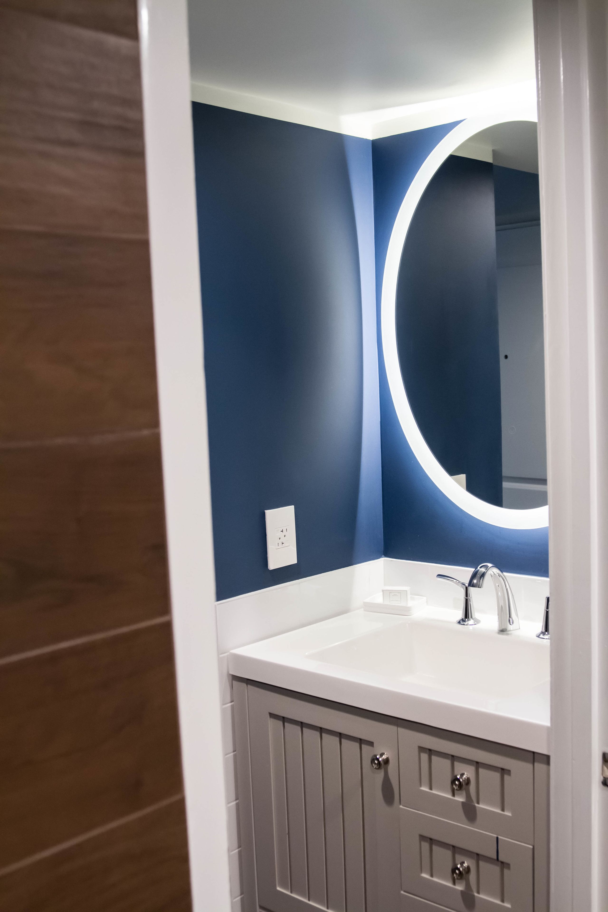 A bathroom with a white sink, a mirror with a circular light, and grey cabinets.