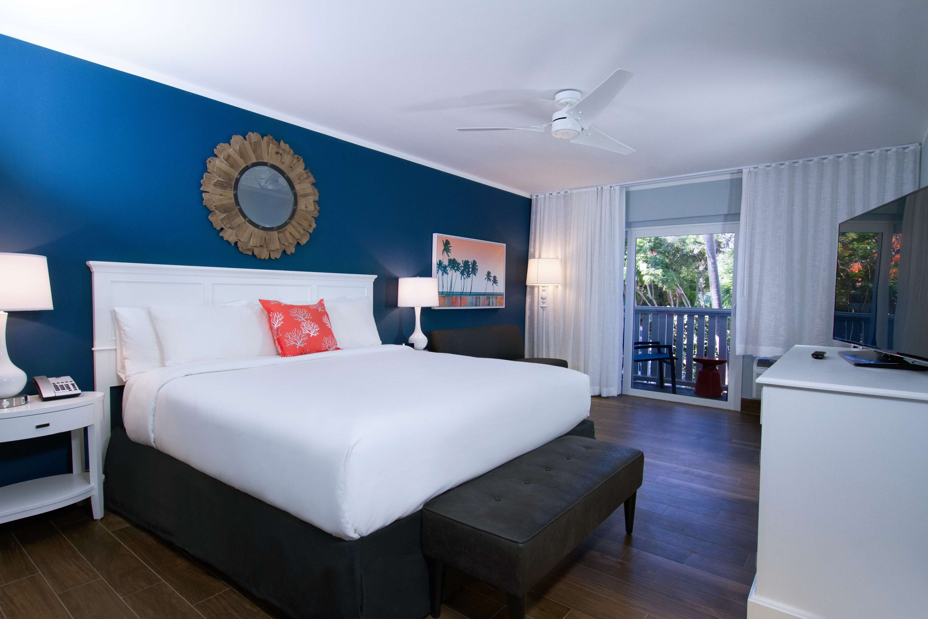 Habitación de hotel con una cama grande, una mesita de noche blanca, un escritorio, un televisor y un balcón con vistas a árboles.
