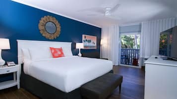 Habitación de hotel con una cama grande, una mesita de noche blanca, un escritorio, un televisor y un balcón con vistas a árboles.
