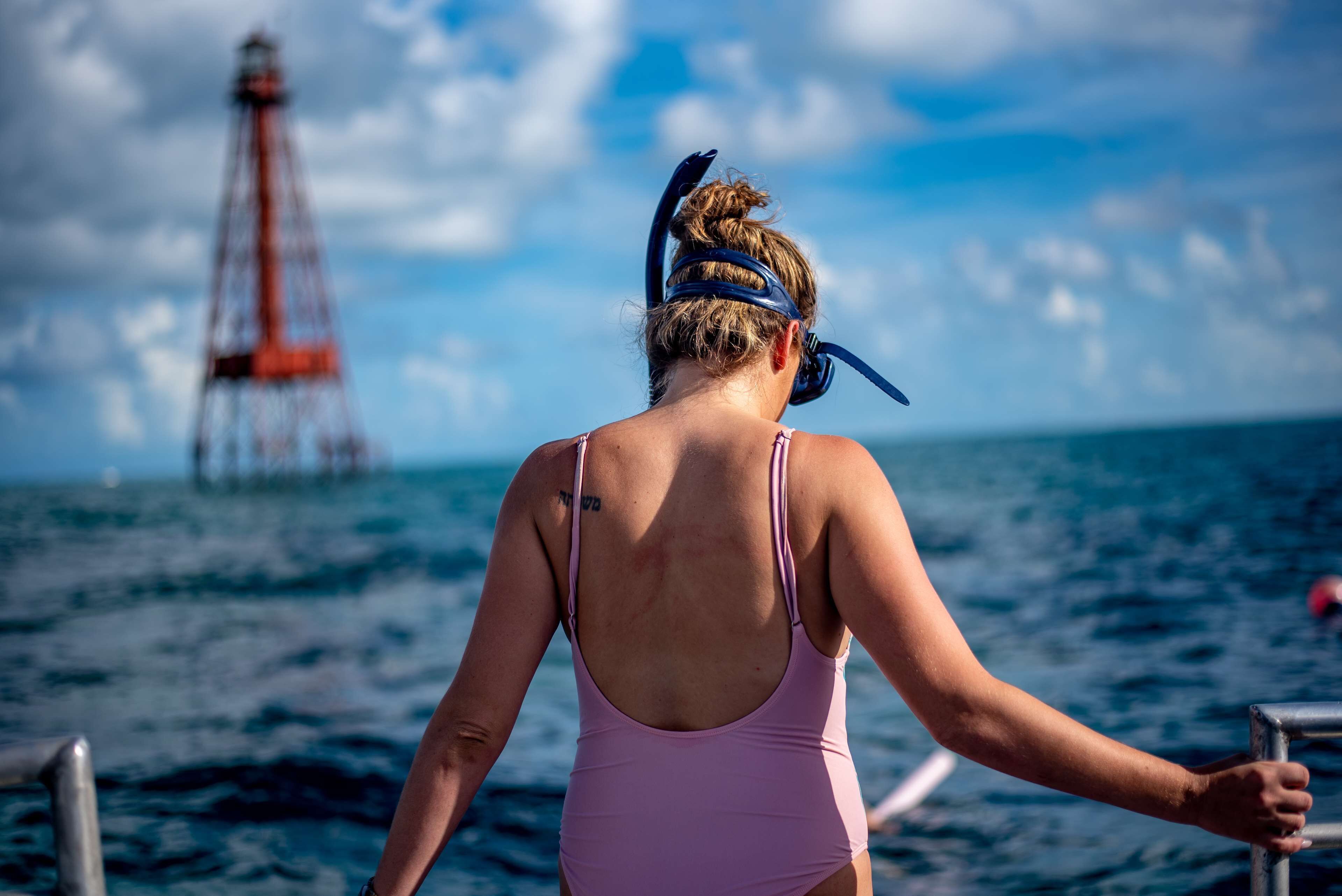 Une personne portant un maillot de bain rose et un bandeau, debout sur un bateau, avec un phare en arrière-plan.