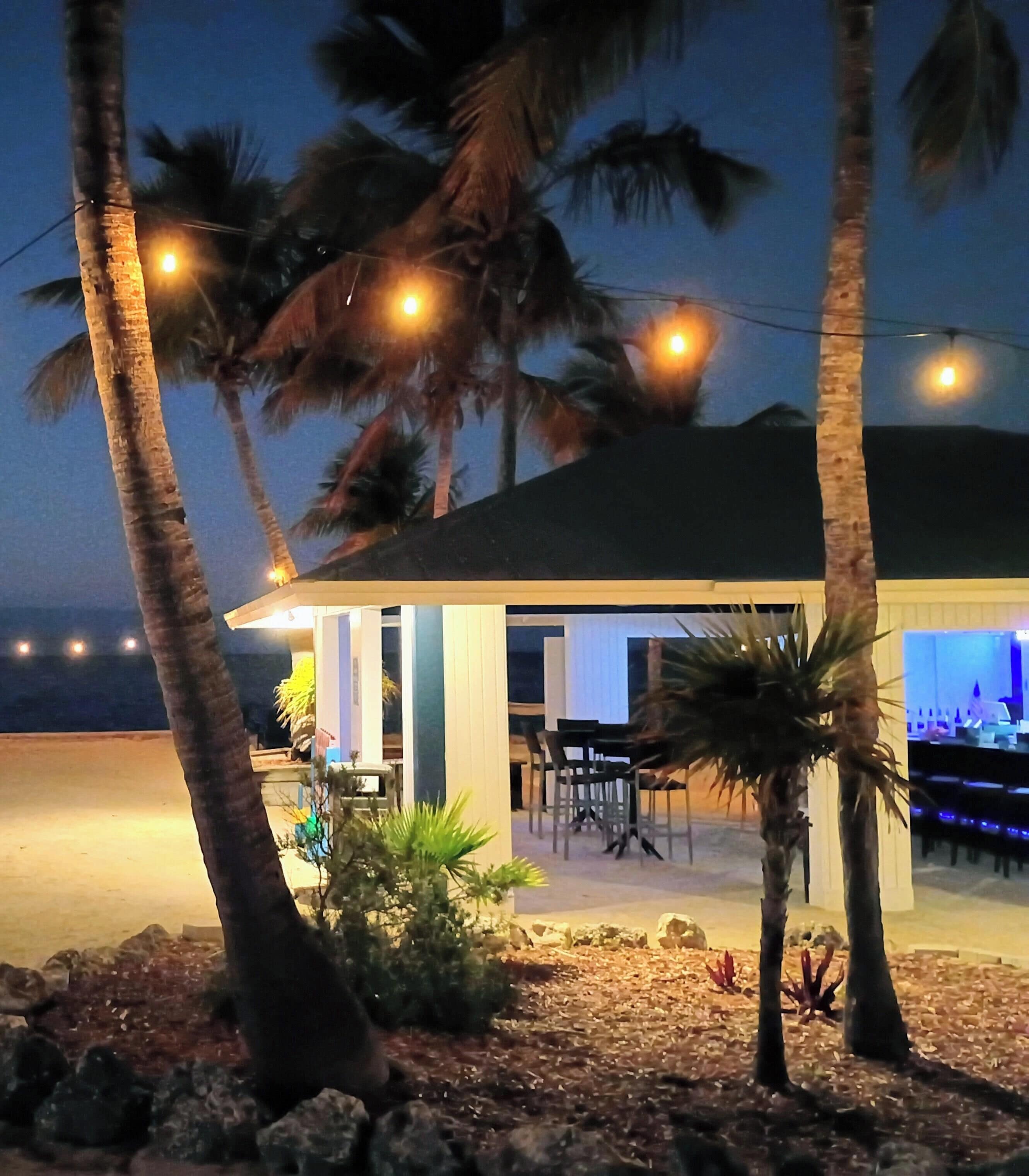 A beachfront property with a dining area, palm trees, and a well-lit exterior.