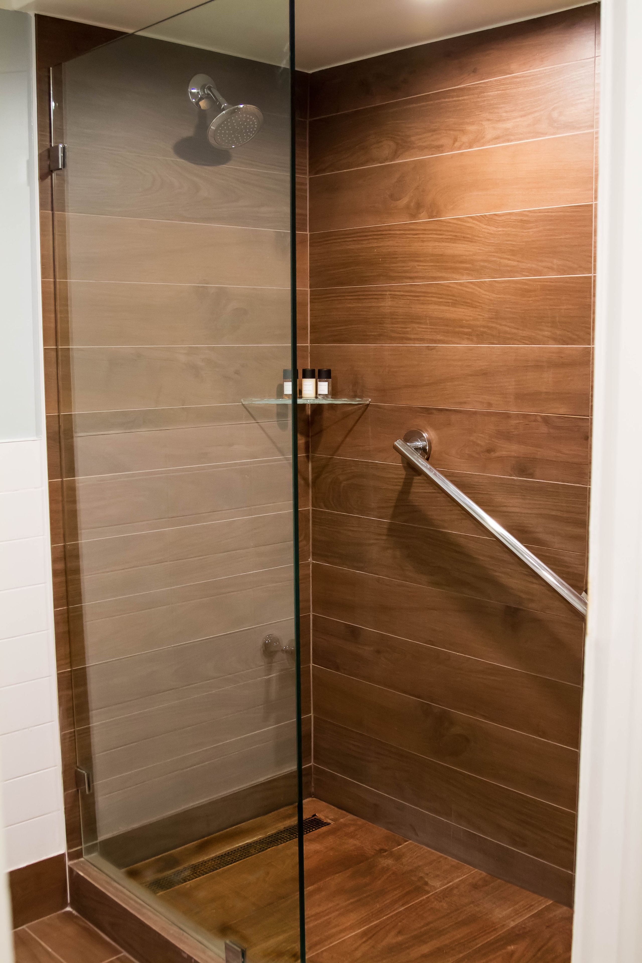 Ducha con cerramiento de vidrio, pared de paneles de madera, repisa de vidrio con artículos de aseo y barra de apoyo metálica.