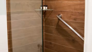 Ducha con cerramiento de vidrio, pared de paneles de madera, repisa de vidrio con artículos de aseo y barra de apoyo metálica.