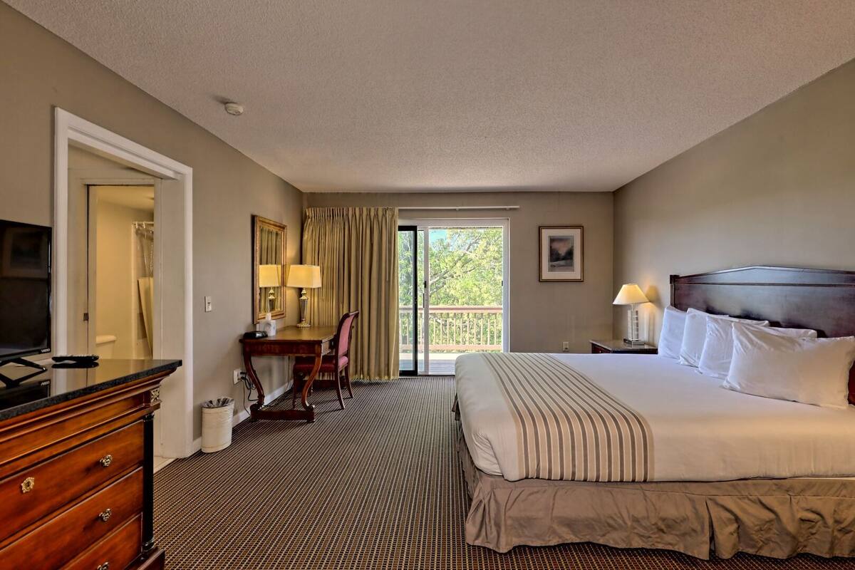 A hotel room with a bed, a desk with a chair, a television, and a balcony with curtains.