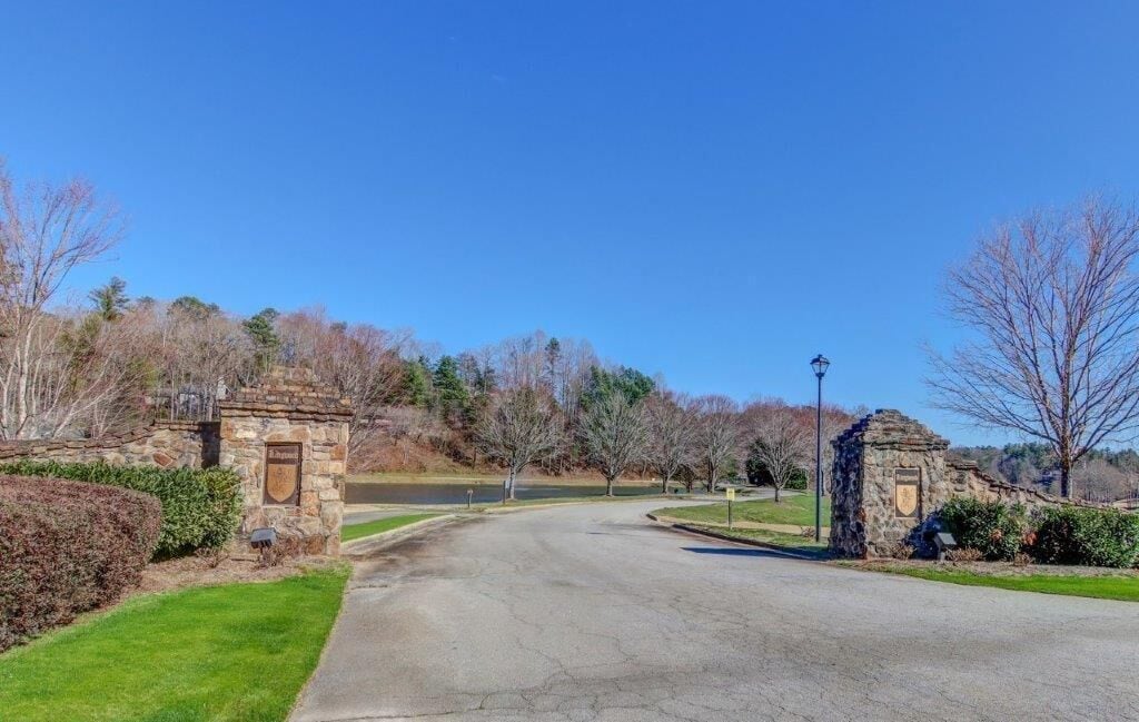 A paved driveway leading to a stone entrance with a plaque, surrounded by trees and a clear sky.