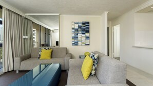 Uma sala de estar moderna com dois sofás cinzentos, um com almofada amarela, e uma mesa de centro de vidro azul. Há uma grande obra de arte abstrata na parede e uma janela com cortinas.
