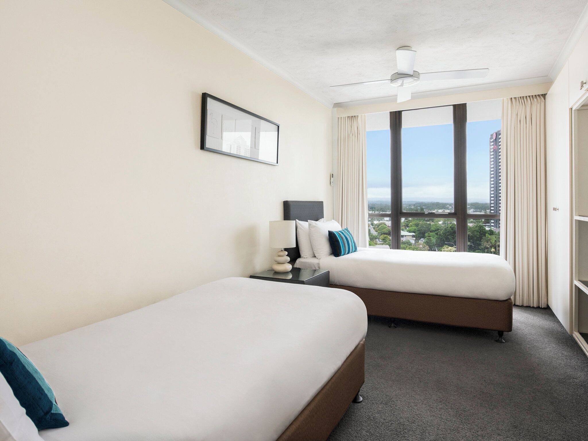 A hotel room with two beds, a ceiling fan, a large window with a city view, and a framed picture on the wall.