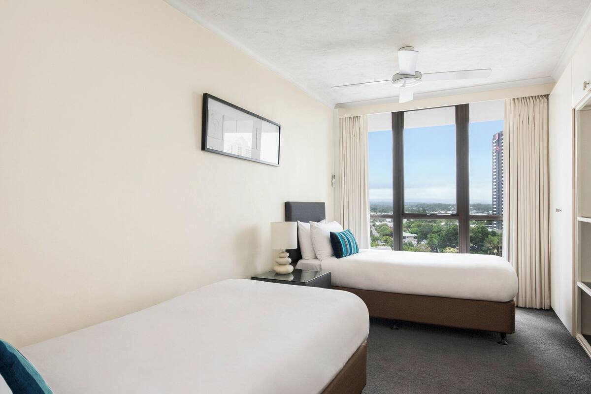 Habitación de hotel con dos camas, un ventilador de techo, un ventanal grande con vistas a la ciudad y un cuadro en la pared.