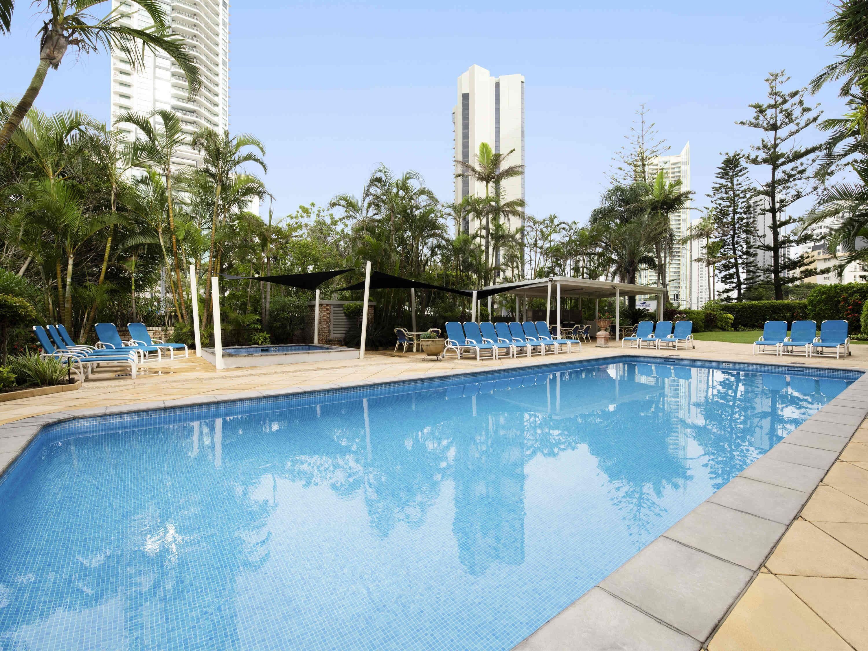 Una piscina con sillas de descanso, rodeada de palmeras y edificios modernos.