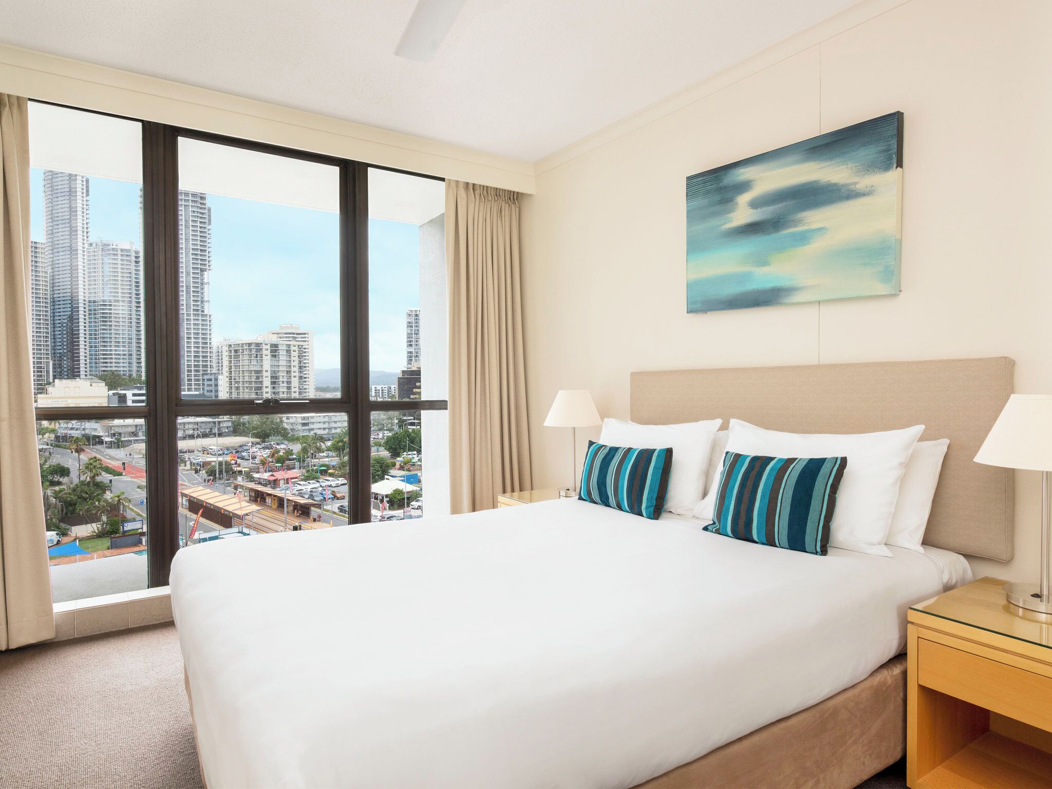 A hotel room with a large bed, two bedside lamps, a city view, and a painting on the wall.