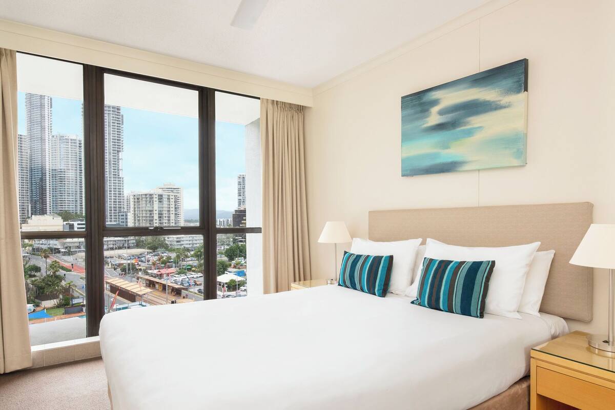 Habitación de hotel con una cama grande, dos lámparas de noche, vista a la ciudad y un cuadro en la pared.