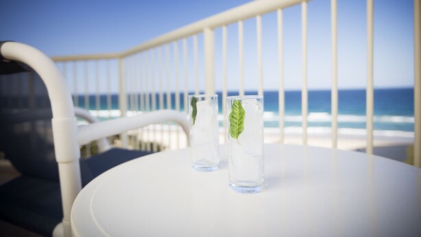 Uma mesa branca com duas taças de vidro com água e uma folha verde, posicionada em frente a um corrimão de varanda com vista para o oceano.