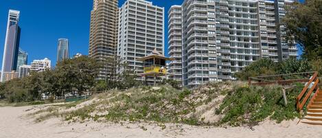 Ein Hotel direkt am Strand, im Hintergrund mehrere hohe Gebäude, ein Sandstrand und eine Holztreppe, die zum Strand hinabführt.