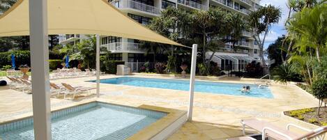 Piscina com espreguiçadeiras, área sombreada e um edifício em vários andares ao fundo.