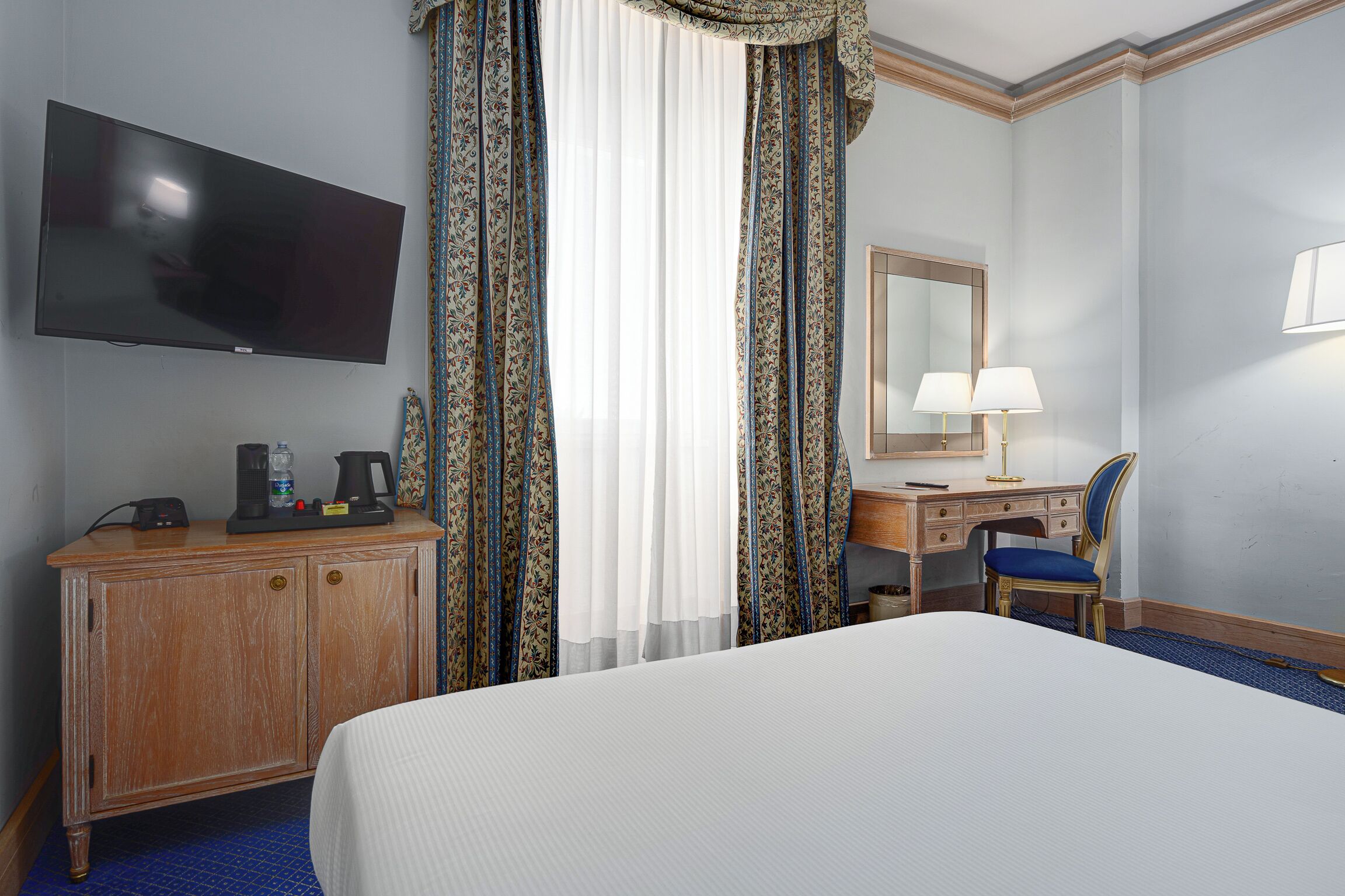 A hotel room with a bed, a desk with a chair, a television, a mirror, and curtains.