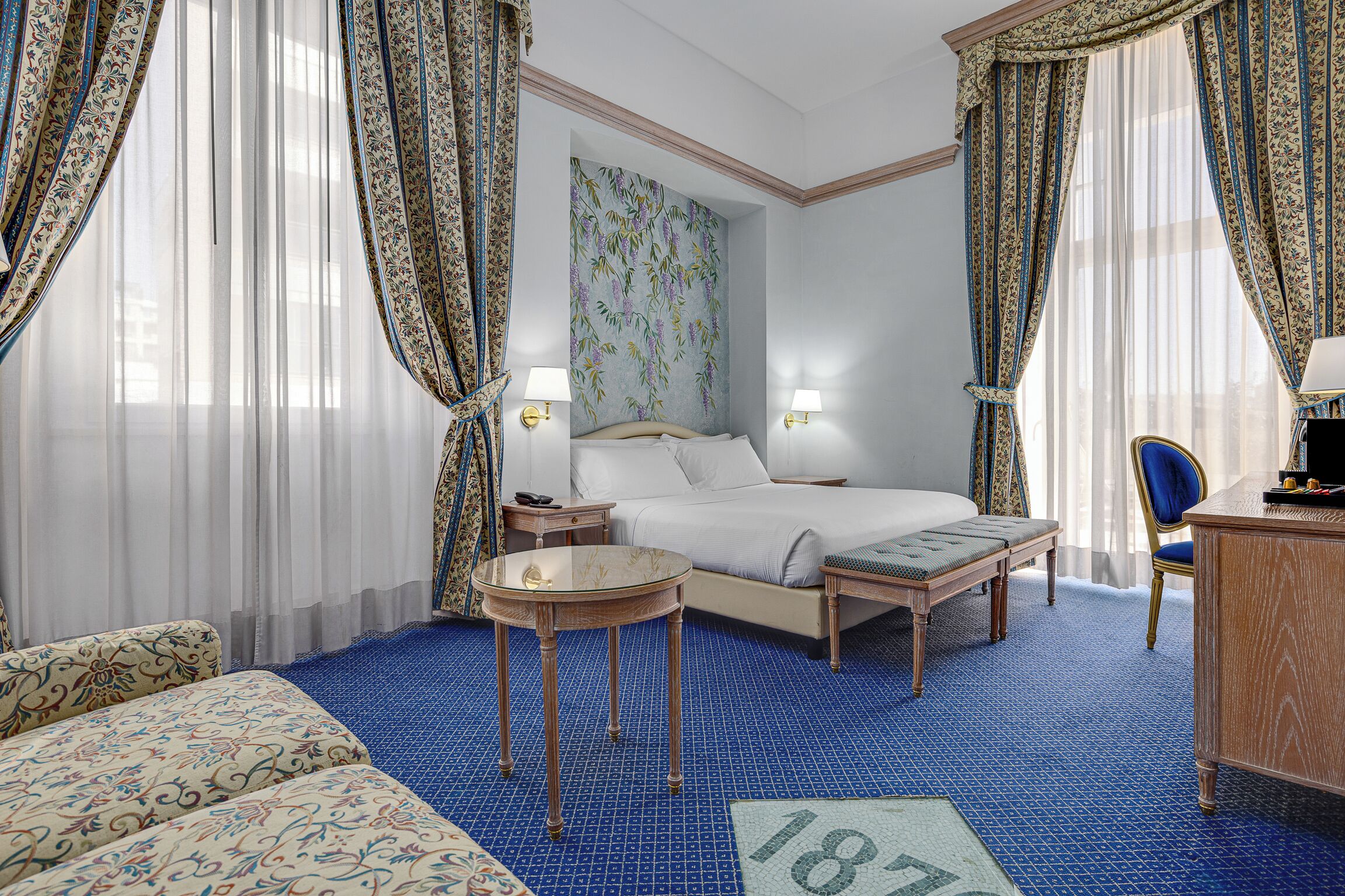 A hotel room with a bed, a desk, a chair, a small table, and a wall with a floral pattern.